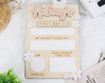 May include: A wooden first day of school sign with the name "Aubrey's" in pink script. The sign includes spaces for "My Favorite Things Are," "I Am," and "When I Grow Up I Want to Be." White daisy accents decorate the sign.