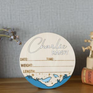 May include: A wooden baby announcement plaque with a blue ocean scene. The plaque has the name "Charlie Randy" written in white script. It has spaces to fill in the date, time, weight, and length of the baby.