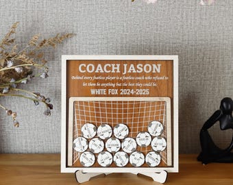 May include: A wooden plaque with a soccer net design and the text "COACH JASON" and "WHITE FOX 2024-2025". The plaque has a white background and a brown wood grain pattern. There are 20 small soccer ball-shaped tokens with names and numbers on them.