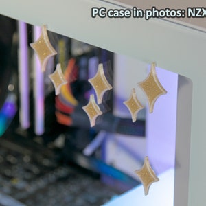 May include: A white PC case with a clear side panel. The case has gold glitter stars on the front and side panels. The text "NZXT H7 Elite (White)" is visible on the top of the case. The text "Tymah's Forge" is visible on the side panel.