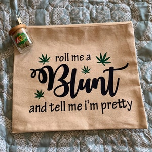 May include: A beige canvas zippered pouch with the text "roll me a Blunt and tell me I'm pretty" in black script and cannabis leaf graphics. A small glass jar with a wooden lid and cannabis inside is next to the pouch.