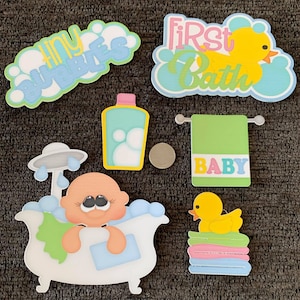 May include: A collection of baby-themed paper cutouts. Includes a baby in a bathtub, a rubber duck, a bottle of baby wash, a towel with "BABY" on it, and text that says "tiny bubbles" and "first bath". The cutouts are in pastel colors.
