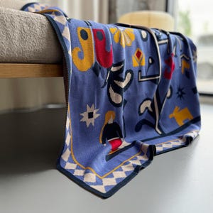 Op de afbeelding: Een blauwe gebreide sprei met een geometrisch en abstract ontwerp. De sprei heeft een rand met een ruitpatroon en diverse vormen in geel, rood en wit. De sprei is over een bank gedrapeerd.