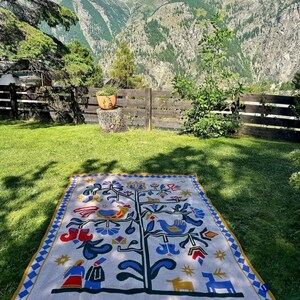 Pu&ograve; includere: Una coperta rettangolare color crema con un disegno dell'albero della vita, con uccelli, fiori e figure in blu, rosso e giallo. La coperta ha un bordo a rombi blu e giallo ed &egrave; appoggiata sull'erba verde.