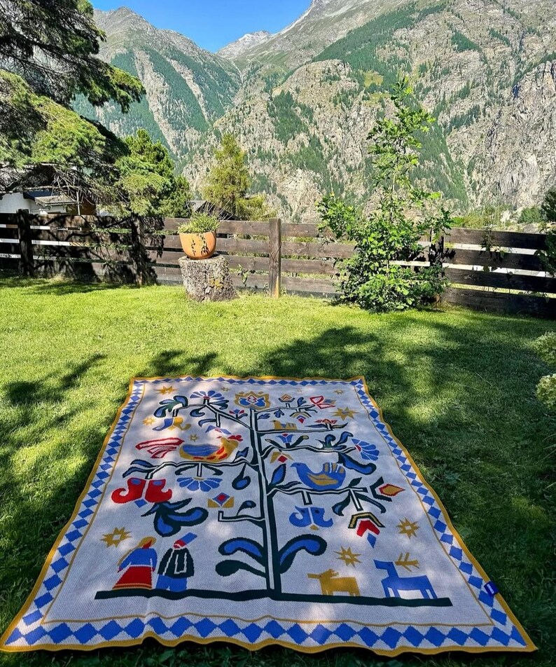 Pu&ograve; includere: Una coperta rettangolare color crema con un disegno dell'albero della vita, con uccelli, fiori e figure in blu, rosso e giallo. La coperta ha un bordo a rombi blu e giallo ed &egrave; appoggiata sull'erba verde.