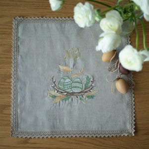 May include: A square, pale grey linen Easter table runner featuring embroidered Easter eggs in a nest, with gold lettering and feathers. The edges are trimmed with a delicate lace border. A vase of white flowers and decorative eggs are in the background.