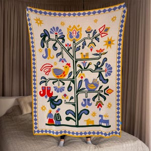 Pu&ograve; includere: Coperta color crema con un disegno dell'albero della vita. L'albero presenta uccelli, fiori e forme geometriche in giallo, blu, rosso e verde. La coperta ha un bordo blu e giallo.