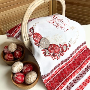 May include: A white Easter basket with a handle, filled with a white embroidered cloth and wooden bowls of decorated Easter eggs. The cloth features red and brown embroidery with floral and egg designs.