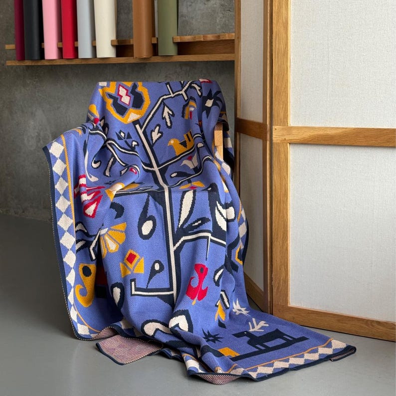 Op de afbeelding: Een blauwe gebreide sprei met een geometrisch en door de natuur ge&iuml;nspireerd ontwerp. De sprei heeft een boomachtig patroon met vogels, herten en abstracte vormen in wit, geel, rood en zwart. De rand heeft een ruitpatroon.