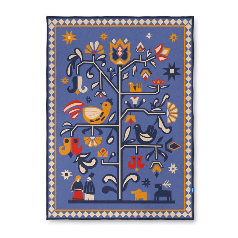 Op de afbeelding: Een blauwe decoratieve deken met een levensboomontwerp. De boom heeft gele, oranje en witte bloemen, vogels en andere volkskuntelementen. De deken heeft een cr&egrave;mekleurige rand met een ruitpatroon.