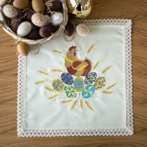 May include: A cream-coloured Easter table runner with an embroidered design. The design features a colourful hen, decorated eggs, and wheat stalks. The runner has a delicate lace trim. A basket of eggs is in the background.