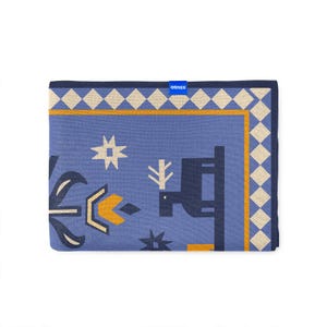 Op de afbeelding: Een opgevouwen blauwe deken met een geometrisch patroon. Het ontwerp bevat vormen in wit, geel en marineblauw. De rand heeft een ruitpatroon in cr&egrave;me en geel. De merknaam "ORNER" is zichtbaar op een blauw label.