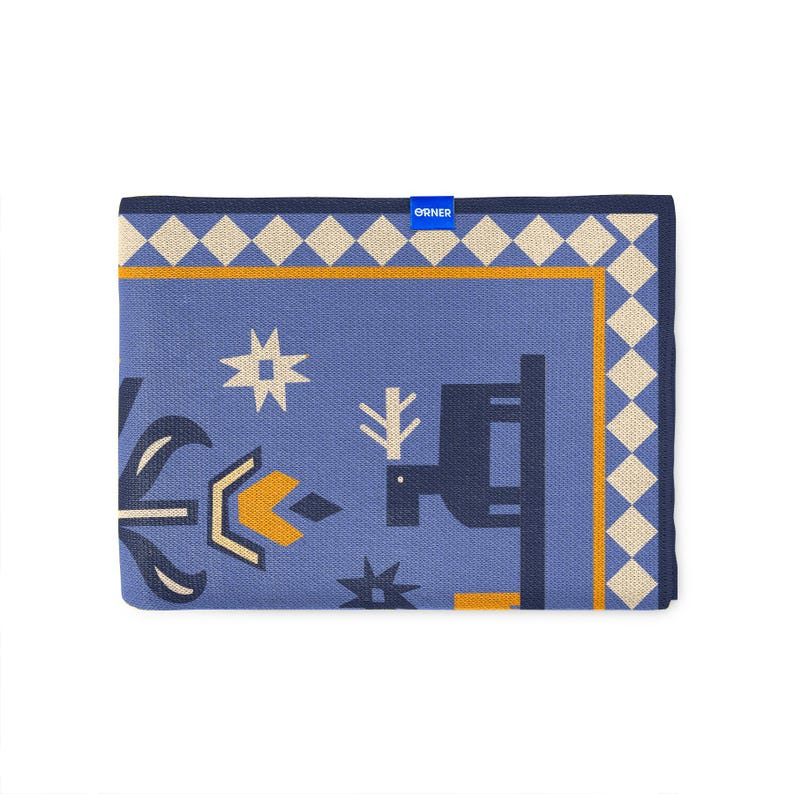 Op de afbeelding: Een opgevouwen blauwe deken met een geometrisch patroon. Het ontwerp bevat vormen in wit, geel en marineblauw. De rand heeft een ruitpatroon in cr&egrave;me en geel. De merknaam "ORNER" is zichtbaar op een blauw label.