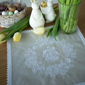 May include: A white embroidered Easter table runner featuring an angel and floral design, edged with lace. Easter decorations, including a basket of eggs, white bunny figurines, and tulips, are arranged around the runner.