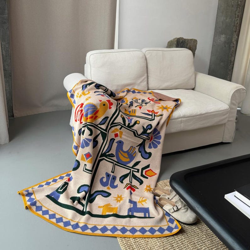 Pu&ograve; includere: Una coperta beige con un colorato design di arte popolare con uccelli, fiori e forme geometriche. La coperta ha un bordo blu e giallo ed &egrave; drappeggiata su un divano bianco. Il design include elementi rossi, blu, gialli e verdi.