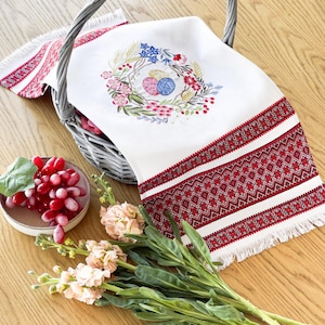 May include: White linen towel with red and white geometric embroidery. A floral wreath with yarn balls decorates the centre. The towel is in a grey wicker basket, with grapes and flowers nearby. The towel has a fringed edge.