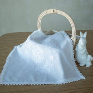 May include: A white embroidered cloth with an angel design and lace trim, draped over a wicker basket with a curved handle. A white ceramic bunny figurine sits beside the basket, creating a peaceful, pastoral scene.