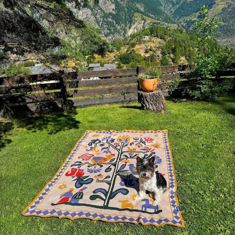 Pu&ograve; includere: Una coperta beige e blu a fantasia, con un disegno di albero e uccelli, &egrave; stesa su un prato verde. Un cagnolino con pelo nero, bianco e marrone &egrave; seduto sulla coperta. La coperta ha un bordo blu e giallo, con una staccionata in legno e montagne sullo sfondo.