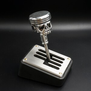 May include: A silver skull-shaped gear shift knob with a piston design. The knob is attached to a black gear shift base with a silver plate and multiple slots.