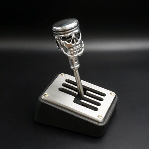 May include: A silver skull-shaped gear shift knob with a black background. The knob is attached to a silver gear shift lever.