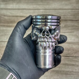 May include: A silver skull-shaped gear shift knob made from a repurposed piston. The knob has a detailed skull design with sharp teeth and a textured surface.