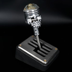 May include: A silver skull-shaped gear shift knob with a detailed design and a silver gear shift lever. The knob is mounted on a black base with a silver gear shift plate.
