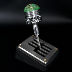 May include: A custom-made gear shifter with a skull-shaped base and a green piston top. The shifter is mounted on a silver metal plate with a black base.
