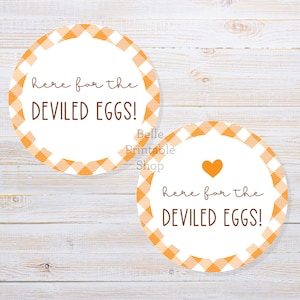 Pode incluir: Dois adesivos redondos com um centro branco e uma borda xadrez laranja e branca. O texto "here for the DEVILED EGGS!" é marrom. Um adesivo tem um coração laranja.