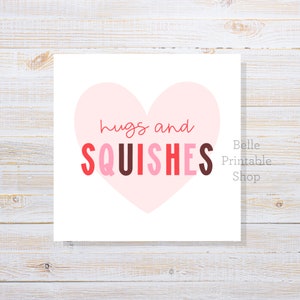 May include: A square print with a light pink heart shape and the words "hugs and SQUISHES" in red, pink, and brown. The words "Belle Printable Shop" are in the bottom right corner. The background is a white wooden surface.