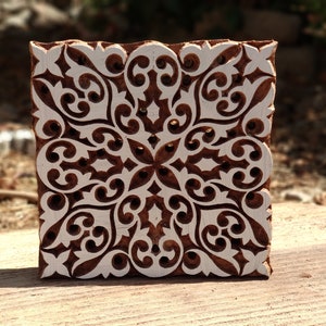 May include: A wooden printing block with a detailed, intricate design carved into it. The design is a symmetrical pattern of swirls and leaves, creating a floral motif. The block is brown with white details.