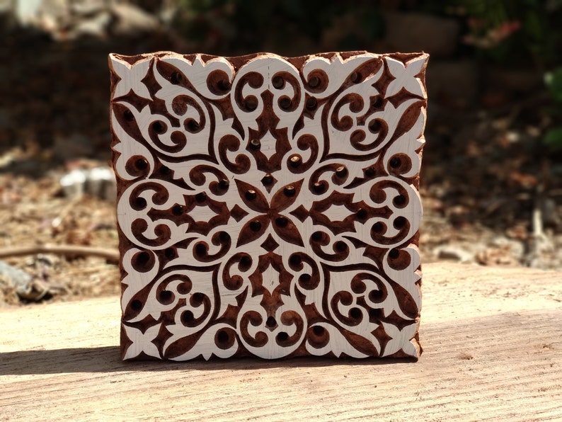 May include: A wooden printing block with a detailed, intricate design carved into it. The design is a symmetrical pattern of swirls and leaves, creating a floral motif. The block is brown with white details.