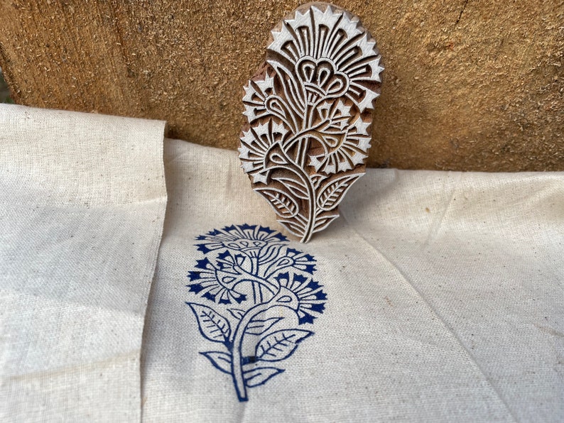May include: A wooden block printing stamp with a floral design. The stamp is used to create a blue floral print on a white fabric.