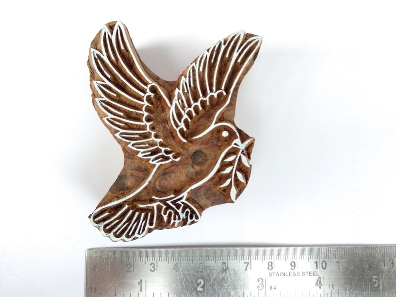 K&ouml;nnte beinhalten: Ein Holzblock-Stempel mit einem wei&szlig;en Design einer Taube, die einen Olivenzweig tr&auml;gt. Der Stempel ist etwa 7,5 cm breit.