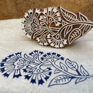 May include: A wooden block printing stamp with a floral design. The stamp is carved with intricate details of flowers, leaves, and stems. The stamp is used to create a blue floral print on fabric.