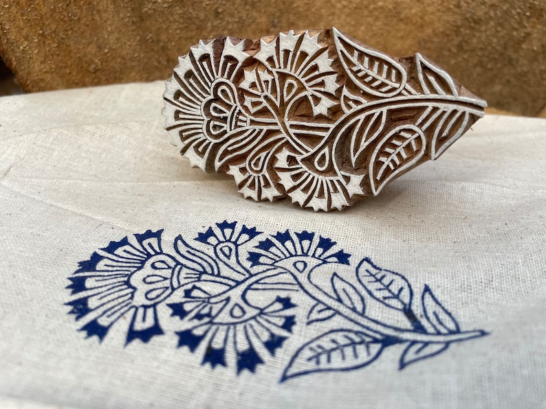 May include: A wooden block printing stamp with a floral design. The stamp is carved with intricate details of flowers, leaves, and stems. The stamp is used to create a blue floral print on fabric.