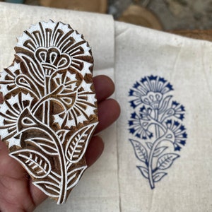 May include: A wooden block printing stamp with a detailed floral design. The stamp is carved with a white floral pattern on a brown wooden background. The stamp is being held in a hand.
