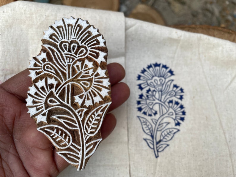 May include: A wooden block printing stamp with a detailed floral design. The stamp is carved with a white floral pattern on a brown wooden background. The stamp is being held in a hand.
