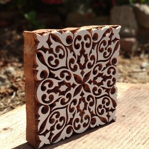 May include: A wooden block printing stamp with a detailed white floral design carved into the surface. The stamp is brown and has a square shape.