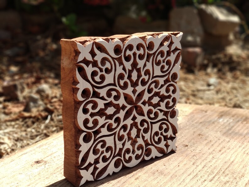May include: A wooden block printing stamp with a detailed white floral design carved into the surface. The stamp is brown and has a square shape.