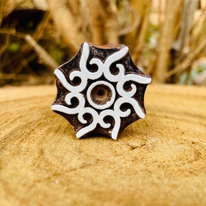 May include: A wooden block printing stamp with a white intricate design. The design features a central circle with swirling patterns radiating outwards.