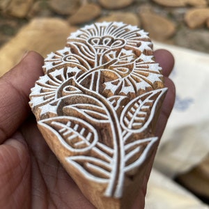 May include: A wooden block printing stamp with a detailed floral design. The design features a large flower with intricate details and leaves. The stamp is carved from light brown wood and has a white design.