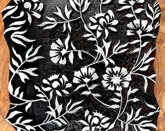 Puede incluir: Un bloque de madera negro con un diseño floral tallado. El diseño presenta flores y hojas blancas sobre un fondo negro.