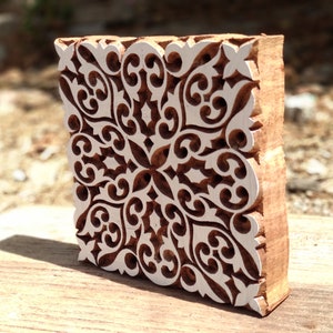 May include: A wooden block printing stamp with a detailed, intricate, white design on a brown background. The design features a repeating pattern of swirls and curves.