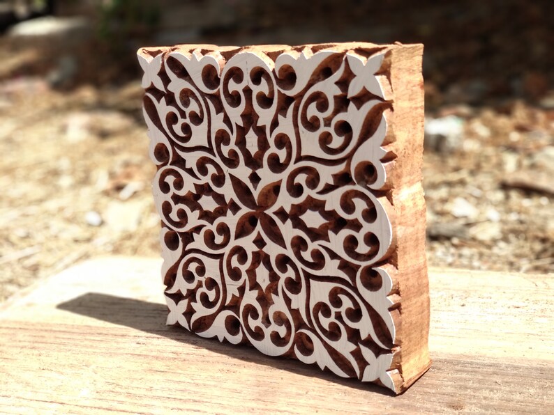 May include: A wooden block printing stamp with a detailed, intricate, white design on a brown background. The design features a repeating pattern of swirls and curves.