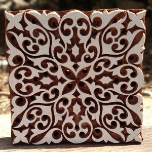 May include: A wooden printing block with a white intricate floral design. The block is square and has a brown color.