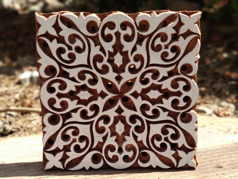 May include: A wooden printing block with a white intricate floral design. The block is square and has a brown color.