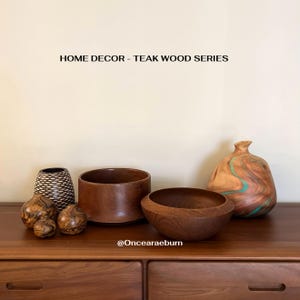 May include: A collection of teak wood home decor items, including bowls, vases, and decorative spheres. The items are various sizes and shapes, with natural wood grain patterns. The text "HOME DECOR - TEAK WOOD SERIES" is displayed at the top.