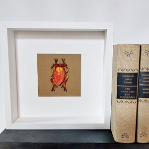 Könnte beinhalten: Gerahmtes Kunstwerk mit einem aufwendig gearbeiteten Insekt aus Perlen, hauptsächlich rot und orange, mit goldenen und braunen Details. Das Kunstwerk ist vor einem braunen Hintergrund in einem weißen Rahmen. Zwei alte Bücher sind zu sehen.