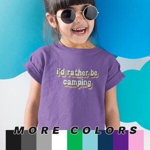 May include: Purple short-sleeved t-shirt with the text "I'd rather be camping" in a retro rainbow font. The shirt is modeled by a young child wearing sunglasses.