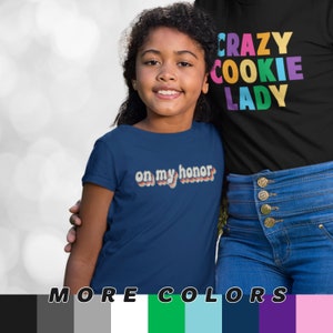 May include: A young person wearing a navy blue t-shirt with the text "on my honor" in a retro rainbow design. The person is standing next to another person wearing a black t-shirt with the text "CRAZY COOKIE LADY" in rainbow colors.